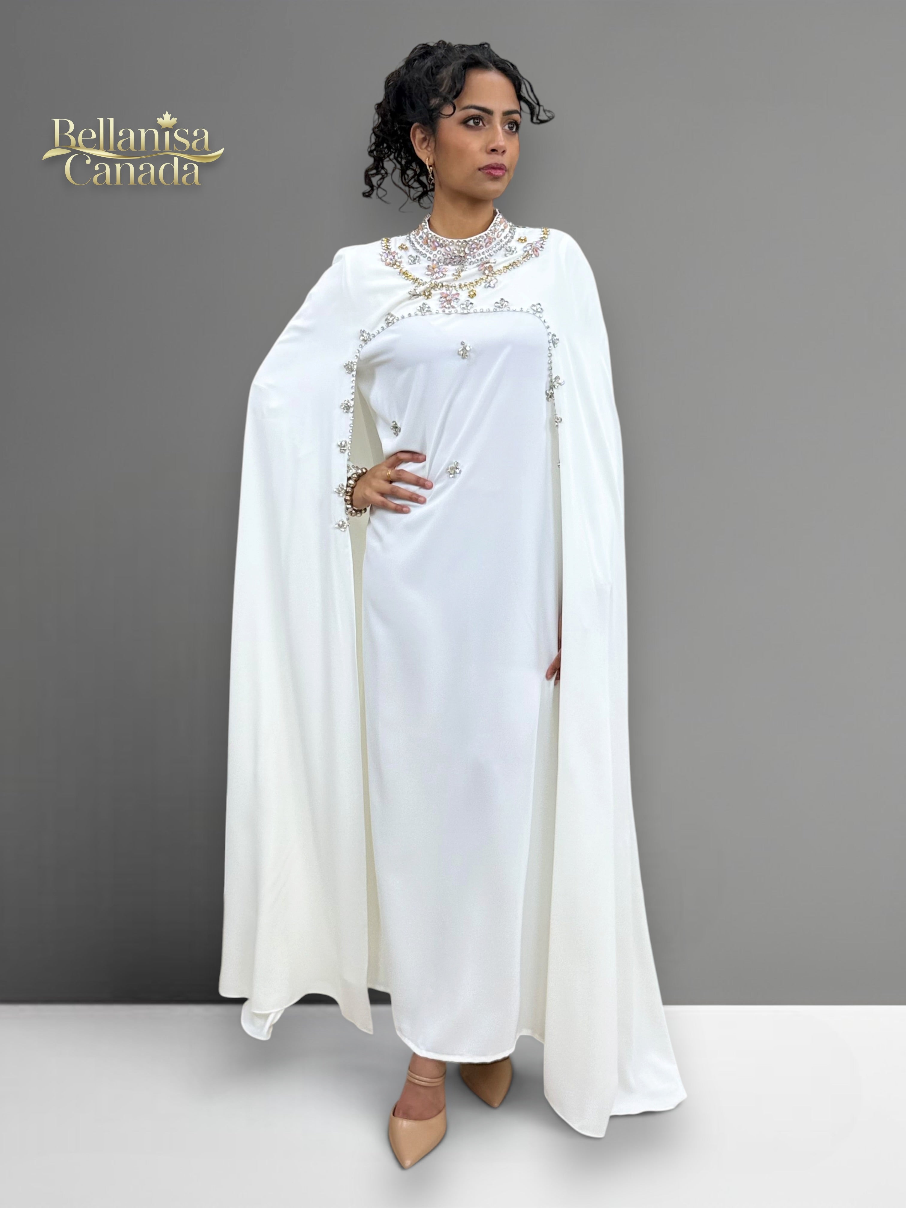 Woman wearing a white dress attached with a cape standing against a gray background. Brand 'Bellanaisa Canada' visible.